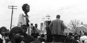 Eine Menschenmenge. Eine Frau ragt heraus, sie blickt in die Ferne. Im Hintergrund ist ein Kirchengebäude