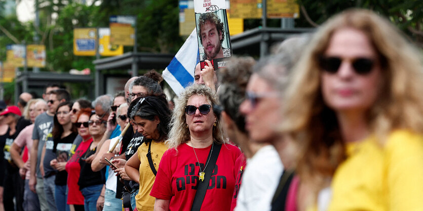 Menschen stehen nebeneinander in einer Reihe, im Hintergrund ist eine Israelfahne zu sehen und ein Foto einer Hamas-Geisel