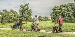 Drei Personen gehen spazieren, sie gehen an Rolatoren, jemamnd sitzt im Rollstuhl