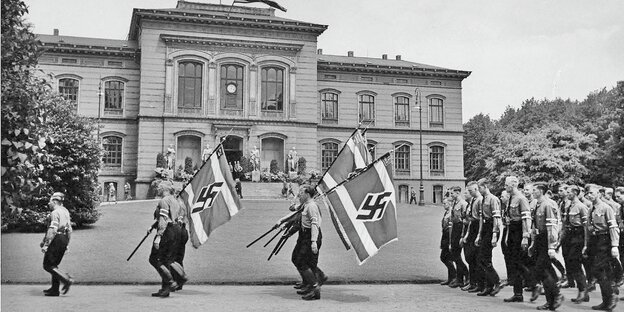 Junge Menschen in Nazi-Uniformen marschieren mit Hakenkreuz-Fahnen vor dem Hauptgebäude der Kieler Universität.