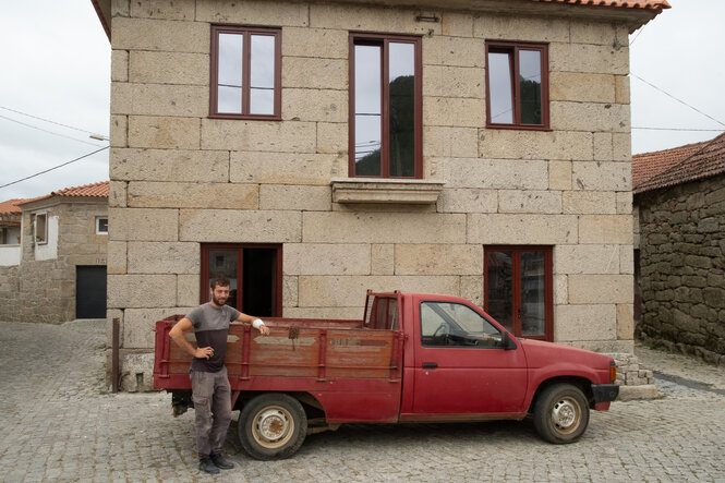 Ein Mann steht, angelehnt an sein Auto, vor seinem Haus