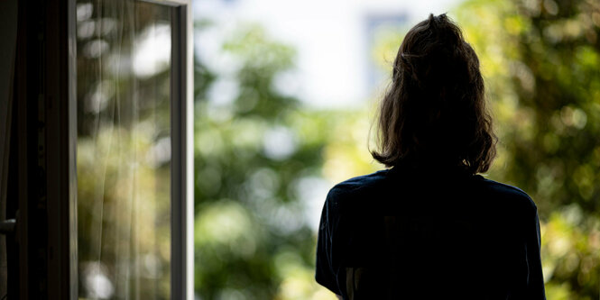 Eine Frau steht mit dem Rücken zum Fenster gewandt und schaut hinaus