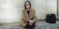 Portrait von Irina Scherbakowa, sitzt mit Brille und braunem Mantel auf einem Steinsockel im Freien vor einem historischen Gebäude