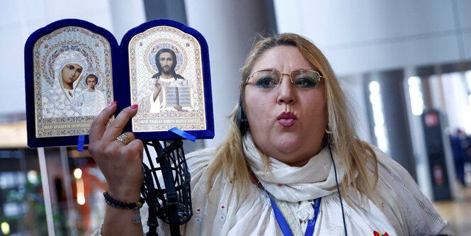 Eine blonde Frau hält eine Aufklapp-Ikone in der Hand in die Kamera. Sie ist aufgeklappt und zeigt Mutter Maria und Jesus.