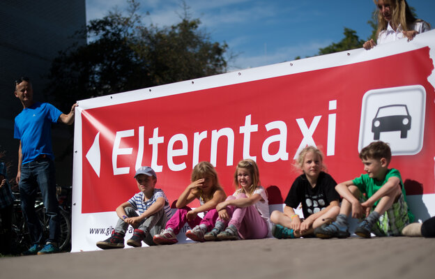 Kinder sitzen auf einer Straße vor einem roten Transparent mit der Aufschrift Elterntaxi