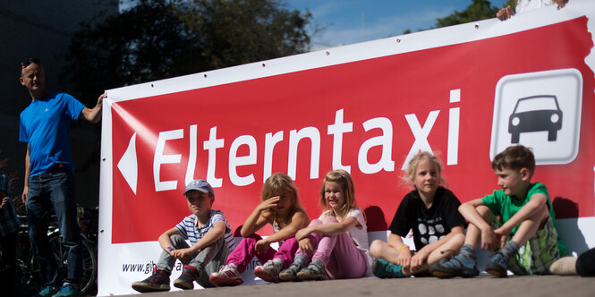 Kinder sitzen auf einer Straße vor einem roten Transparent mit der Aufschrift Elterntaxi