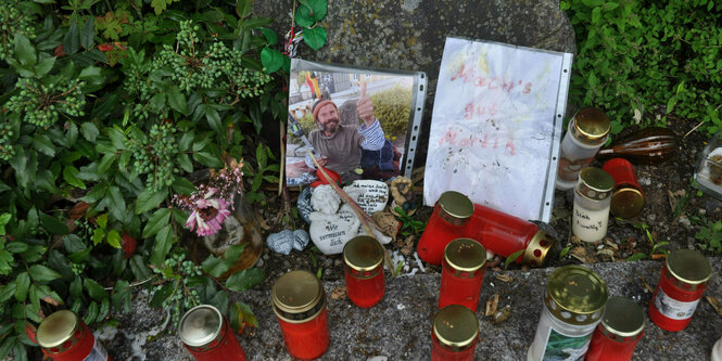 Grabkerzen stehen im Grünen. In der Mitte das Foto eines bärtigen Mannes , der lächelt und den Daumen einer Hand der Kamera entgegenstreckt