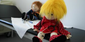 Zwei Handpuppen mit Stetkoskop sitzen auf einer Liege in einem ärztlichen Untersuchungszimmer