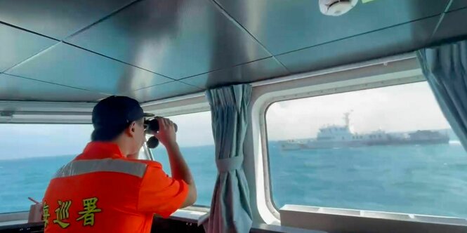 Ein Mann in einem orangefarbenen T-Shirt beobachtet aus dem Fenster eines Schiffs ein anderes Schiff mit dem Fernglas