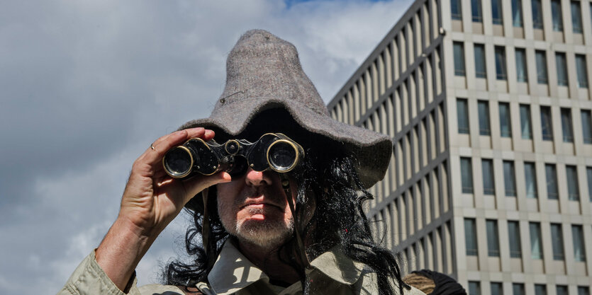 Mann mit Schlapphut und Fernglas vor modernem Bürogebäude