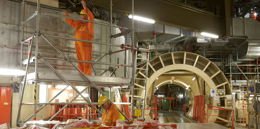 zwei Männer in orangenen Overalls, einer auf Gerüst, Turbine im hintergrund