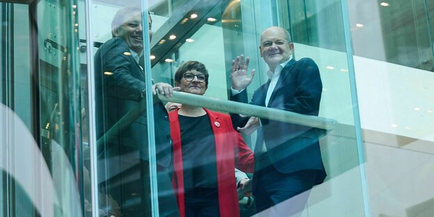 Saskia Esken im roten Mantel und Lars Klingbeil mit Olaf Scholz stehen winkend in einem fahrenden gläsernen Fahrstuhl