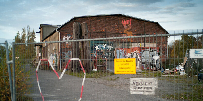 eine Backsteinhalle, dabvor ein Bauzaun, an dem Schilder hängen: "Betreten verboten" und "Vorsicht Ratten"