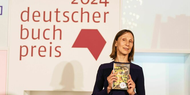 Martina Hefter vor einer Tafel „2024 Deutscher Buchpreis“, ein Exemplar ihres Romans hochhaltend