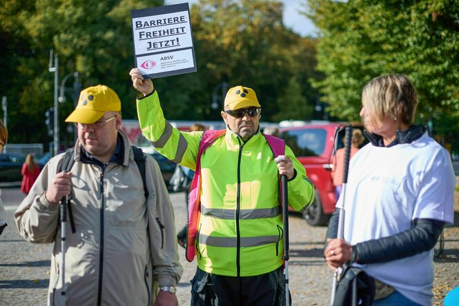 Drei sehbehinderte Menschen mit Taststöcken auf einer Protestaktion, zwei Männer tragen ein gelbes Basecap mit den drei schwarrzen Punkten,