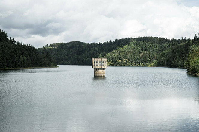 Wasseroberfläche von bewaldeten Hügeln umsäumt, in der Mitte steht ein Wasserturm