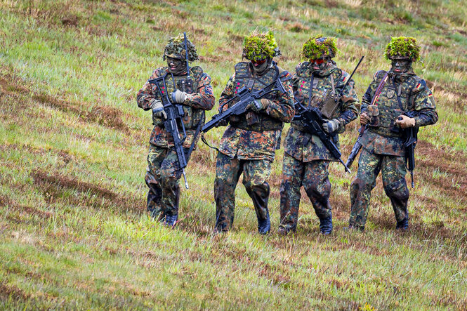 Vier Soldaten in Tarnuniform bewegen sich hintereinander über eine Wiese