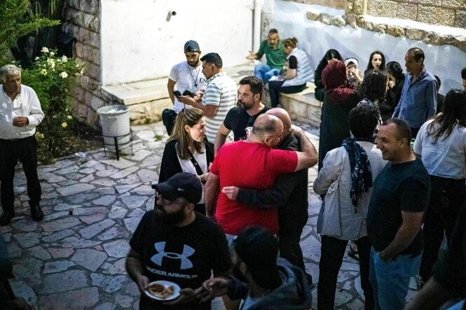 Männer und Frauen stehen in einem Hof, sprechen, lachen, umarmen sich, essen und trinken