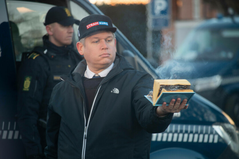 Rasmus Paludan steht auf der Straße, hinter ihm ein Polizist und er hält einen Koran in der ausgestreckten Hand, der vor sich hinkokelt