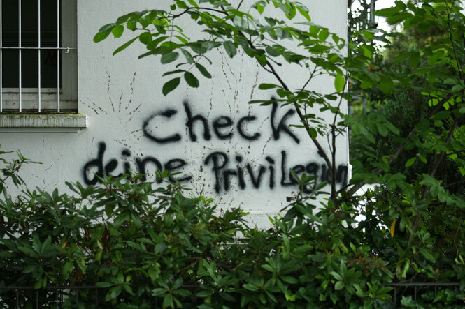 An einer Hauswand, vor der grüne Büsche wuchern steht: Check deine Privilegien