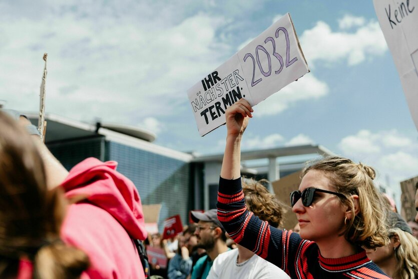 Protestaktion von Therapeut*innen, eine junge Frau mit Sonnenbrille hält ein kleines Schild hoch: Ihr nächster Termin: 2032