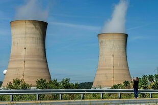 Kühltürme von Three Mile Island vor blauem Himmel, von Bäumen und Palmen umgeben. Eine Frau fotografiert