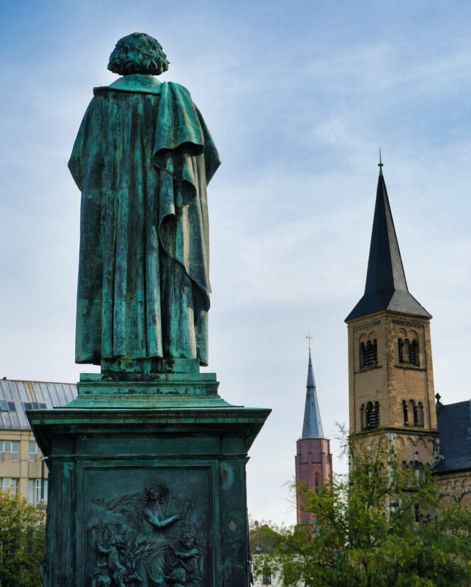 Eine Beethovenstatue von hinten vor Kirchtürmen.