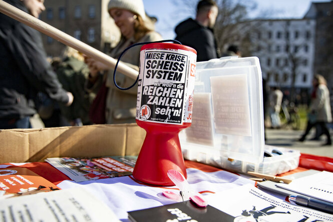 Eine Spendebox mit dem Schrift "Miese Krise, scheiss System, wir wollen die Reichen zahlen sehen", steht auf einem Stand während des Internationalen Frauentages am Oranienplatz in Berlin am 8. März 2024