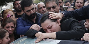 Eine Frau trauert über einem Sarg, Männer stehen neben ihr