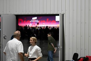 Ines Schwerdtner unterhält sich vor einer geöffneten Tür vor dem Saal mit einem Delegierten auf dem Bundesparteitag der Partei Die Linke in Halle/Saale. Hinter der Tür sieht man den Slogan "Die Linke".