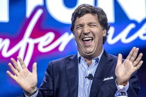 Tucker Carlson hebt die Hände und lacht überzogen vor einem Neonschild.