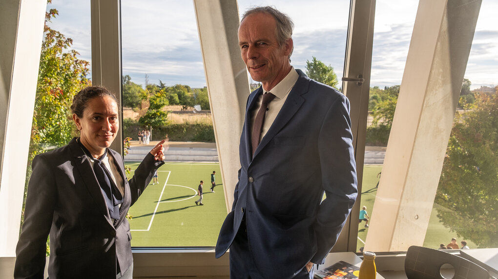 Eine Frau und ein Mann stehen an einem Fenster, sie zeigt auf ein Fussballfeld