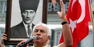 Ein Mann hält ein Mikrofon und hebt einen Arm in die Höhe, im Hintergrund die türkische Flagge und ein Bild von einem Mann