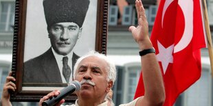 Ein Mann hält ein Mikrofon und hebt einen Arm in die Höhe, im Hintergrund die türkische Flagge und ein Bild von einem Mann