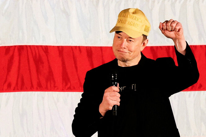 Elon Musk trägt eine goldene "Make America Great Again" Baseballcap, mit Mikrofon in der rechten Hand am Körper und der linken Faust erhoben vor einem weiß-rot-weißen Hintergrund, welcer zu einer großen US Flagge gehört.