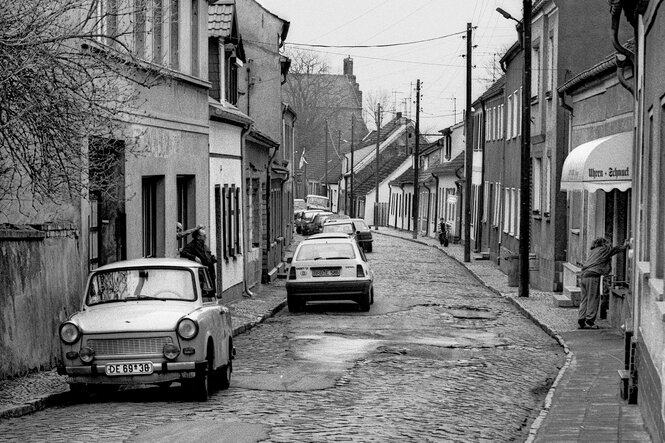 Blick in eine holprige Straße mit kleinen Häusern, am Straßenrand parken Trabbis und dieersten Westautos