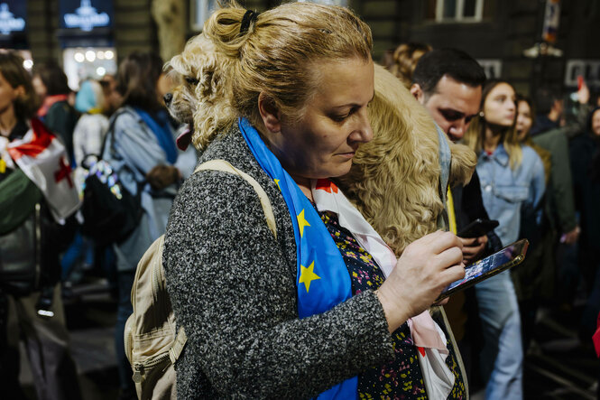 Eine Frau steht in einer Menschenmenge und schaut auf ihr Smartphone, sie trägt eine europäische Fahne um den Hals, auf ihrer Schulter sitzt ein Hund