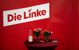 Das Bild zeigt eine Tisch mit Blumen und einer Flasche vor einem Stellwand mit dem Schriftzug "Die Linke"