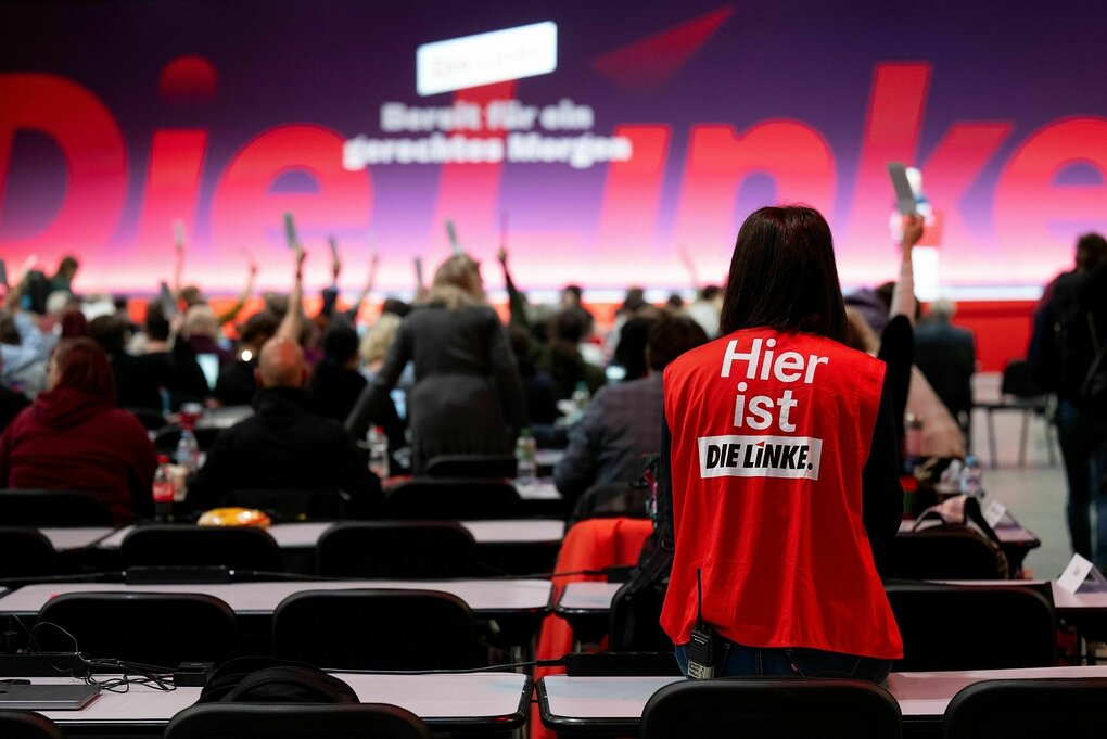 Person mit Weste von Linkspartei bei Parteitag vor Tischreihen