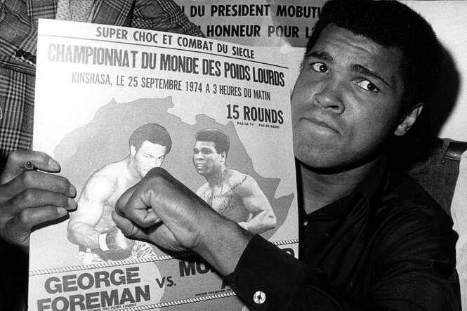 Schwarz-weiß Aufnahme von Boxer Muhammad Ali wie er ein Werbeplakat für seinen Kampf gegen George Foreman in der Hand hält