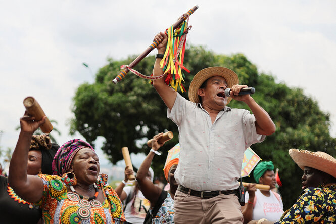 Menschen aus verschiedenen indigenen und afro-kolumbianischen ethnischen Gruppen singen, halten ihre instrumente nach oben,imVordergrund ein Mann mit Hut und Mikrofon