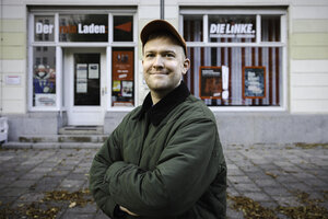 Porträt von Paul Uebler, er steht mit Basecap lächelnd vor dem Roten Laden - Parteibüro mit Plakaten und zwei Schaufenstern