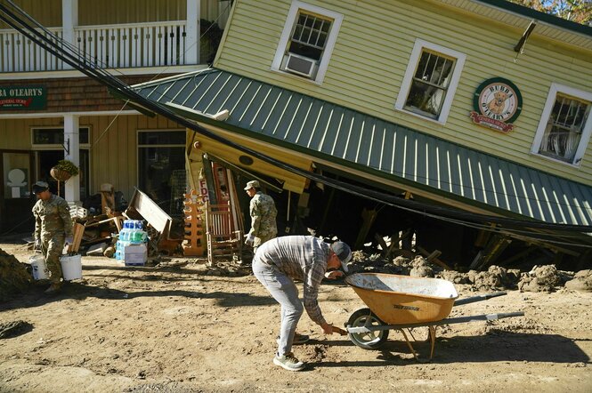 Männer mit und ohne Unifrom bei Aufräumarbeiten nach dem Hurrican, einer fixiert eine Schubkarre ein anderer trägt Eimer, im Hintergrund ein Haus, das schief an der Straße steht, das Untergeschoss ist teilweise weggebrochen