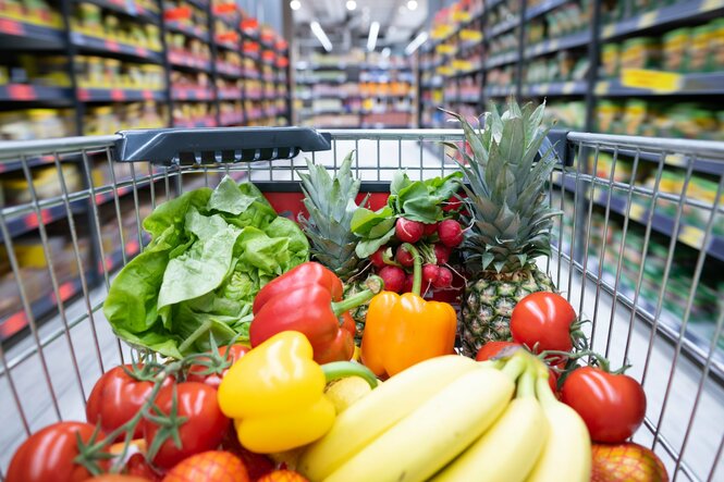 Mit Obst ud Gemüse gefüllter Einkaufswagen in einem Supermarkt-Gang.