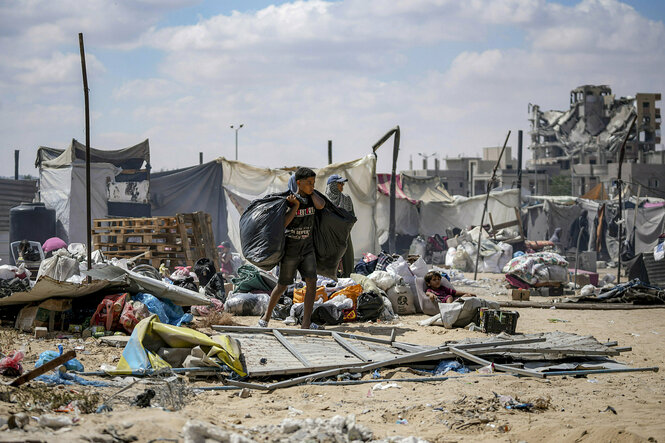 Ein Junge, beladen mit schwarzen Tüten, geht in Gaza über einen Paltz auf dem selbstgebaute Zelte stehen, die Häuser im Hintergrund sind zerbombt