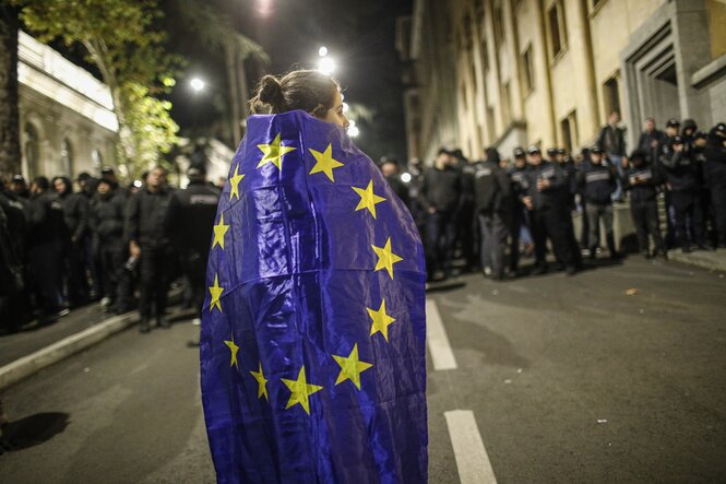 Eine in eine EU-Fahne gewandte Person steht einer Reihe von Uniformierten in der Dunkelheit auf einer Straße gegenüber