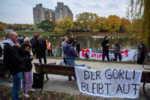 Ein Dutzend Leute steht in einem Park am Wasser. An einer Bank ist ein Transparent angebracht mit der Aufschrift 