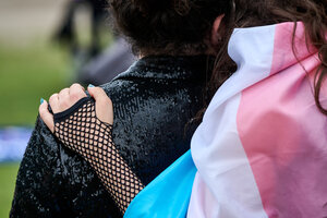 Eine Person mit trans Flagge um die Schultern umarmt eine andere Person von hinten