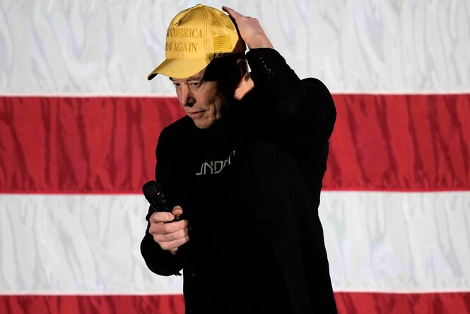 Elon Musk steht vor einer US-Fahne, er trägt eine goldene Basecap, auf die die Worte "Make America great again" gestickt sind