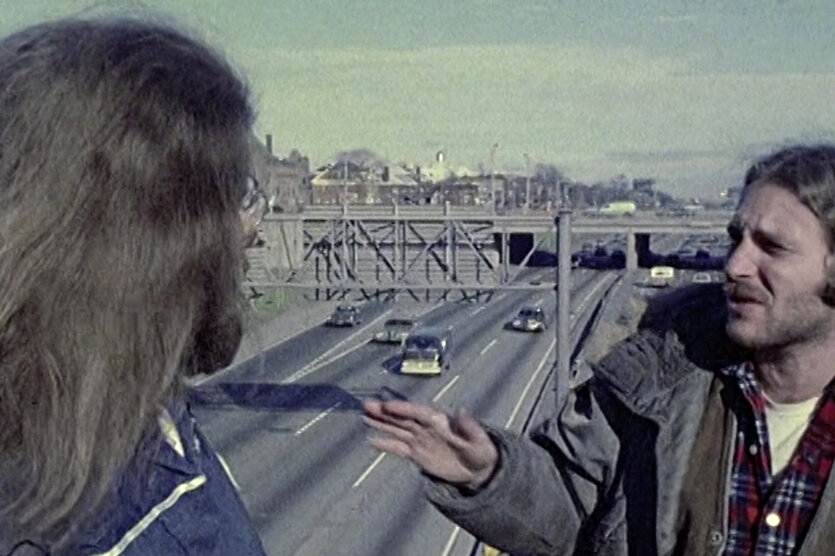 Still aus einem Film von Robert Kramer, zwei langhaarige, weiße Männer stehen auf einer Autobrücke, hinter ihnen eine US-amerikanische Stadt